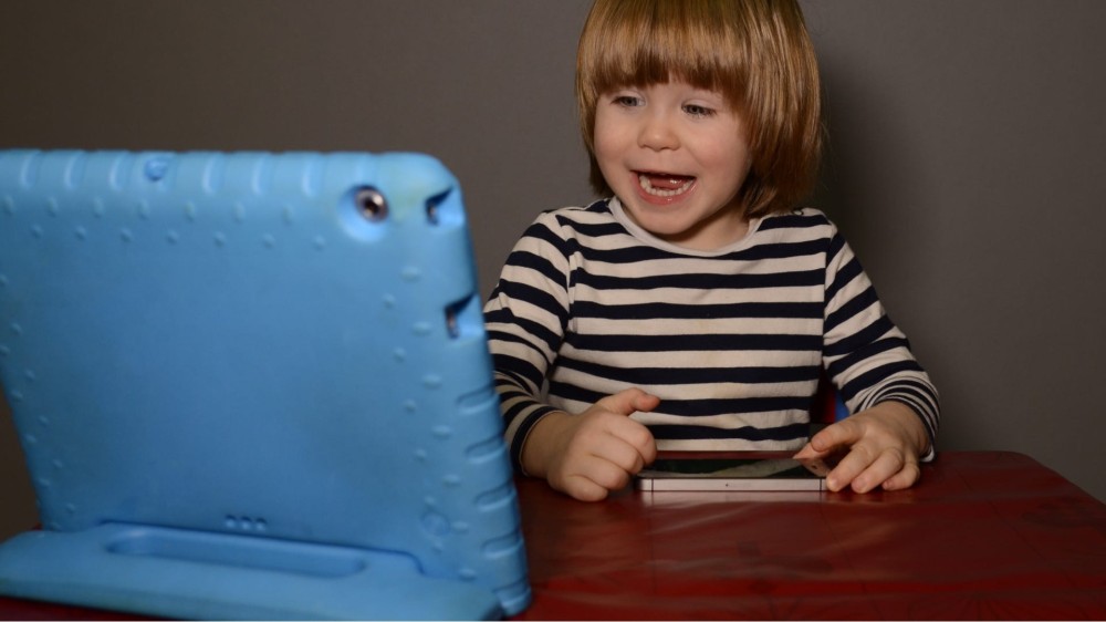 A young boy sits at a table watching something on a tablet in a blue standing case.