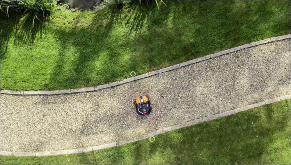 A Worx Landroid Vision smart lawn mower navigatin over a pathway to mow the other side of a backyard.